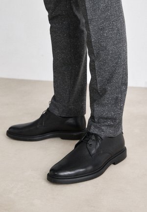 Schwarze Lederschuhe mit glatter Textur, runder Zehenform und Schnürdesign, kombiniert mit dunkelgrauen gemusterten Hosen.