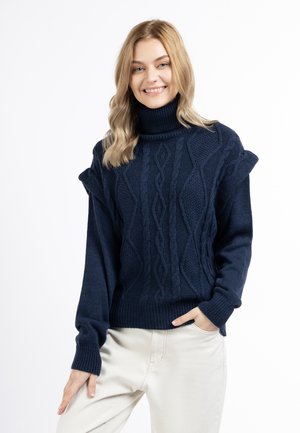 Donna sorridente con capelli biondi indossa un maglione a collo alto blu navy con trecce e pantaloni chiari, in piedi con una mano in tasca.