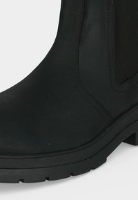 Gros plan d'une botte cheville en cuir noir avec une semelle épaisse en caoutchouc et un panneau latéral élastique.