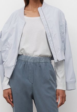 Femme portant une veste bomber gris clair sur un t-shirt blanc à manches longues et un pantalon ample bleu-gris à taille élastique, debout devant un fond uni.