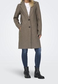 Manteau marron en mélange de laine avec un revers cranté, deux boutons et des poches latérales, porté avec un jean bleu et des bottines noires.