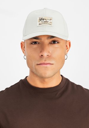 Vīrietis ar baltu cepuri ar taisnstūra formas logotipa plāksteri un brūnu krekliņu ar apaļu kaklu, skatās taisni pretī baltā fona priekšā.