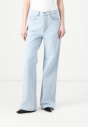 Persona con vaqueros azul claro de pierna ancha, top blanco y zapatos negros de punta, de pie sobre un fondo blanco liso.
