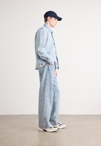 Chaqueta de mezclilla azul clara y jeans de pierna ancha, combinados con deportivas blancas y marinas. El modelo está de lado frente a un fondo neutro.