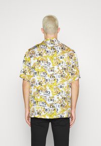 Mies, jolla on lyhyet vaaleat hiukset, pukeutuneena valkoiseen lyhythihaiseen paitaan, jossa on keltaisia barokkikuvioita ja mustaa tekstiä, yhdistettynä mustiin housuihin.