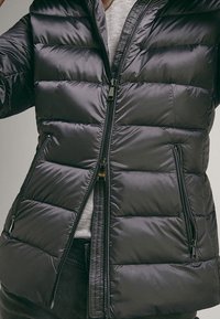 Giacca a vento nera con zip frontale, caratterizzata da sezioni trapuntate orizzontali, tasche laterali e una consistenza morbida e lucida.