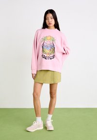 Pink oversized sweatshirt with a beach graphic, paired with a green skirt and light pink sneakers. Smooth fabric and relaxed fit.