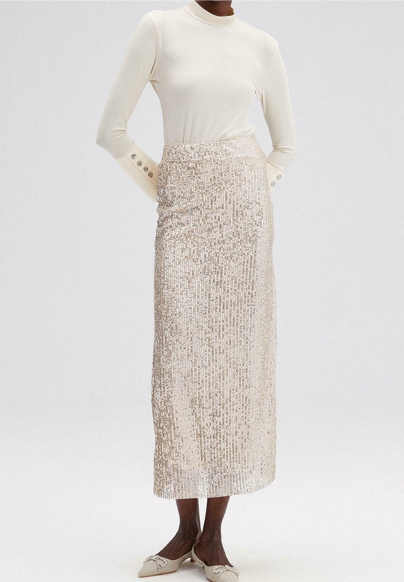Femme portant un haut à manches longues crème, une jupe midi argentée à sequins taille haute et des chaussures assorties à talons bas pointus.