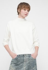 Pullover branco de malha com gola alta, punhos e bainha canelados. Combinado com uma saia de denim em padrão de pele de cobra, nas cores azul claro e verde.
