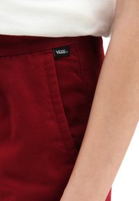 Pantaloni bordeaux realizzati in tessuto testurizzato, con tasche laterali e un piccolo etichetta nera "VANS" sulla vita. Vista ravvicinata del lato.