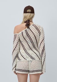 Top all'uncinetto con design a maglia aperta, strisce crema e marrone, stile a spalla unica. Abbinato a pantaloni corti coordinati e un cappellino da baseball marrone.