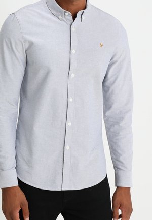 Hombre que lleva una camisa de manga larga azul claro con botones y un pequeño logo bordado en el lado izquierdo del pecho.