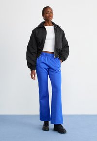 Black bomber jacket with a collar, white cropped shirt, and bright blue flared pants. Model wearing black lace-up boots.
