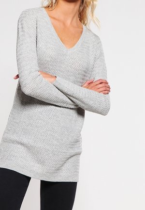 Kobieta ubrana w jasno szary, teksturowany sweter z dekoltem w serek i długimi rękawami, krzyżująca ramiona, w czarnych spodniach na jednolitym tle.