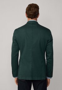 Blazer vert foncé avec une coupe ajustée, fente simple et manches longues. Dispose de deux boutons, revers crantés et une texture lisse et tissée.