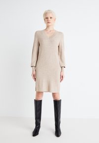 Abito beige a scollo a V con maniche lunghe, texture a costine e spacchi laterali, abbinato a stivali in pelle nera alti con punte appuntite.