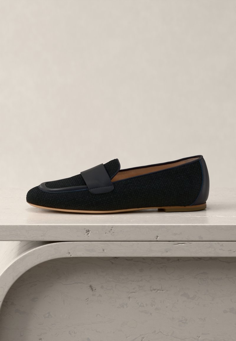 Μαύρο πλεκτό δερμάτινο μοκασίνι με επίπεδη ταμπά σόλα και δερμάτινη λεπτομέρεια λουριού, τοποθετημένο σε ανοιχτόχρωμη υφή πέτρινης επιφάνειας.