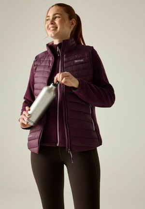 Mujer joven con chaleco y chaqueta de exterior morados, sosteniendo una botella de agua plateada y sonriendo frente a un fondo liso.