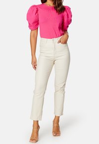 Top de manga abullonada rosa combinado con jeans ajustados crema y tacones de tira transparente. El conjunto presenta un diseño casual pero moderno.