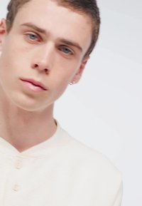 Prenda de color beige claro con una superficie texturizada, presenta un cuello redondo y un cierre de botones, mostrada en un modelo con cabello corto y un arete.