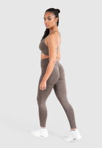 Completo sportivo in tessuto marrone composto da reggiseno e leggings, caratterizzato da un design aderente, una texture senza cuciture e una lieve lucentezza, abbinato a scarpe da ginnastica bianche.