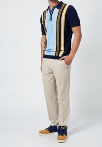 Man in een kortlijf polo met blauwe, gele en zwarte verticale strepen, beige broek en blauwe en tan sneakers, staand met een hand in zijn zak.