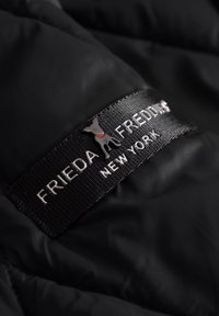 Zwart gewatteerd stof met een gestructureerd oppervlak, voorzien van een gestikte label met "FRIEDA FREDDI" en een klein grijs hondenicoon.