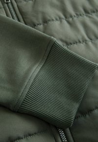 Gros plan sur la manche d'une veste vert olive montrant un poignet en maille côtelée, un tissu matelassé et une fermeture éclair métallique partiellement visible.