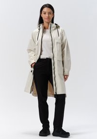 Giacca leggera beige con cappuccio, due tasche frontali e dettagli neri, abbinata a pantaloni neri e sneakers nere. Tessuto liscio.