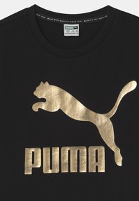 Mustad musta puuvillane T-särk, millel on metallik kullavärvi Puma logo ja tekst julges kirjas, ümmargune kaelus, sile tekstuur.