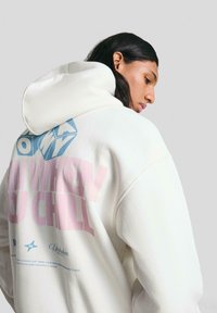 Felpa bianca con grafica testuale blu e rosa sulla schiena. Materiale morbido; grande cappuccio con una vestibilità comoda. Il testo distintivo recita: “CHILL.”
