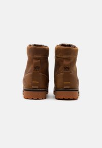 Une paire de bottes en cuir marron avec des cols rembourrés et des semelles robustes, présentée de dos sur un fond blanc.