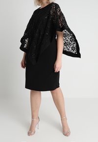 Frau trägt ein schwarzes knielanges Kleid mit einem durchsichtigen Spitzen-Poncho und silbernen High-Heels, steht vor einem schlichten Hintergrund.