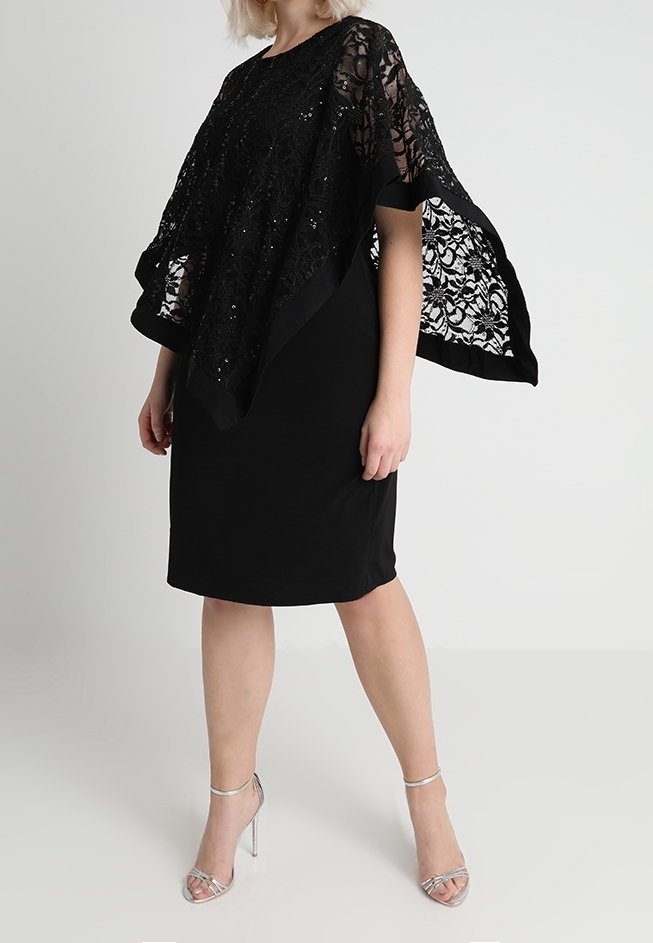 Frau trägt ein schwarzes knielanges Kleid mit einem durchsichtigen Spitzen-Poncho und silbernen High-Heels, steht vor einem schlichten Hintergrund.