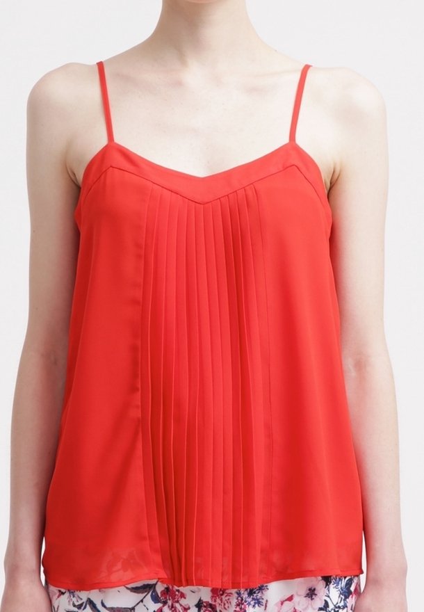 Femme portant un haut rouge sans manches avec de fines bretelles et des plis verticaux sur un pantalon à motif floral.