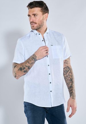 Kurzärmliges weißes Leinenhemd mit Button-Down-Kragen, Brusttasche und schwarzen Knöpfen. Getragen mit dunkelblauen Jeans.