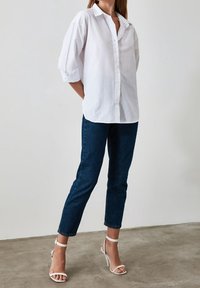 Blusa blanca de botones con mangas enrolladas, combinada con jeans ajustados azul oscuro y sandalias de tacón blancas. Tejido suave y corte casual.