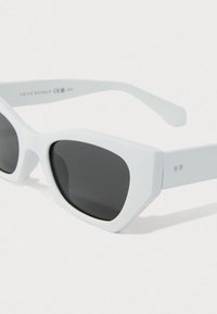 Bílé sluneční brýle ve tvaru kočičích očí s širokými obroučkami a tmavými skleněnými čočkami, umístěné na jednobarevném bílém pozadí.