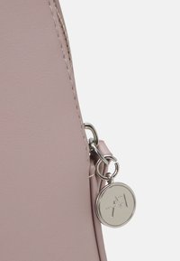 Primer plano de la esquina de un bolso de cuero malva con cremallera plateada y colgante redondo de metal con letras estilizadas "A7" en relieve.