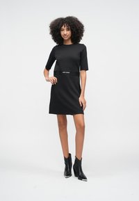 Černé krátké šaty s krátkým rukávem a přiléhavým pasem, doplněné kontrastním páskem s logem. Kombinované s elegantními černými kotníkovými botami. Hladká tkanina.