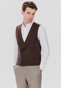 Brązowa kamizelka podwójnie zapinana, wykonana z teksturowanej tkaniny, z dużymi guzikami i kołnierzem z wycięciem, noszona na białej koszuli i w paskowanych spodniach.