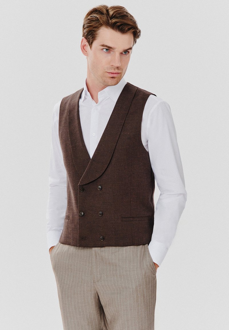 Brązowa kamizelka podwójnie zapinana, wykonana z teksturowanej tkaniny, z dużymi guzikami i kołnierzem z wycięciem, noszona na białej koszuli i w paskowanych spodniach.