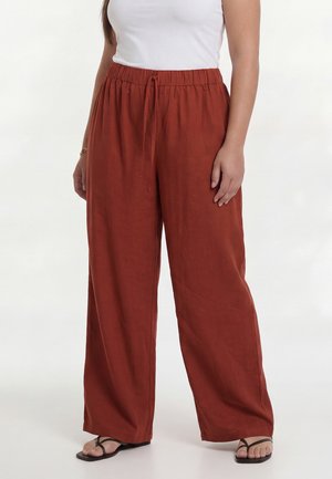 Kvinde iført vidde rustfarvede bukser med elastisk talje og snøre, parret med sorte sandaler med stropper.