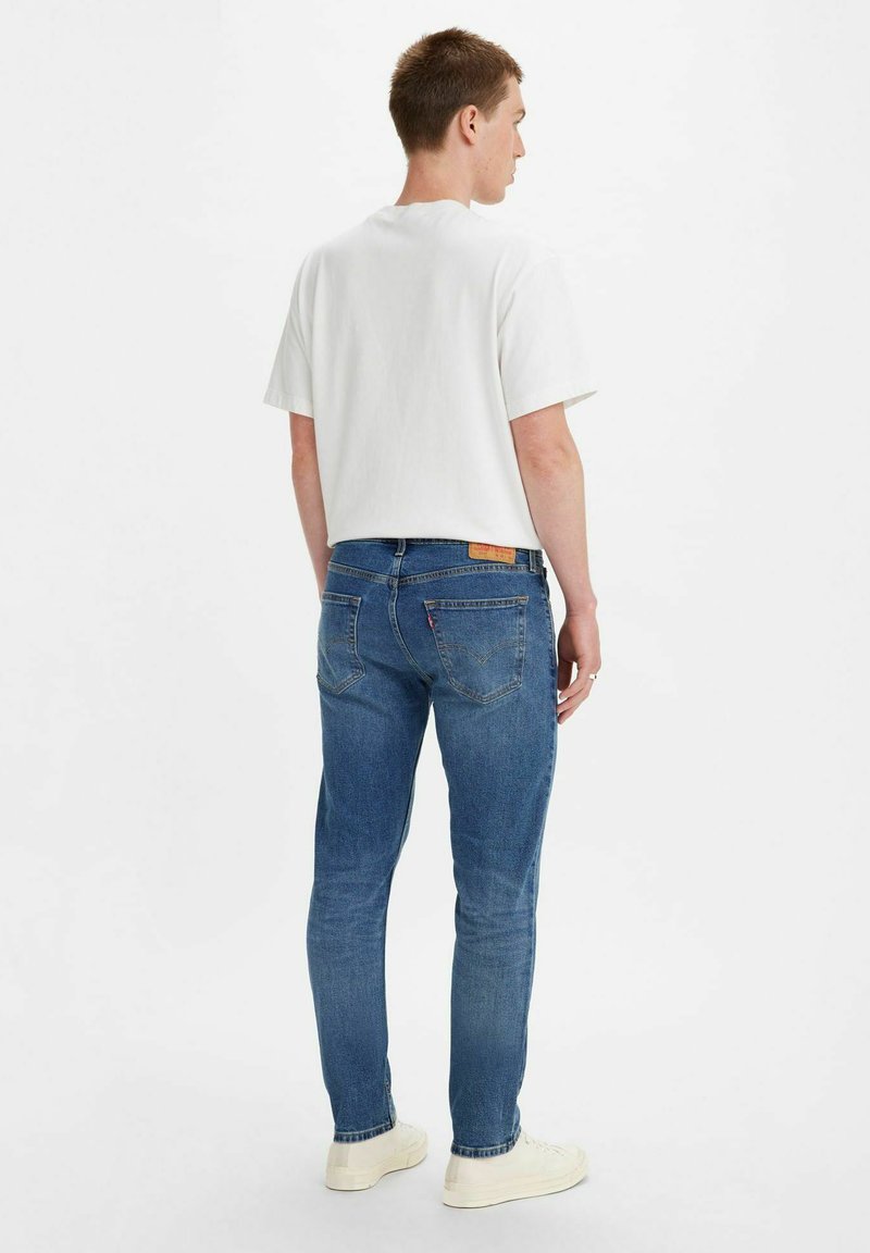 T-shirt bianco a maniche corte abbinato a jeans blu in denim; i jeans presentano tasche posteriori e un'etichetta beige sulla vita. Indossato con sneaker bianche.