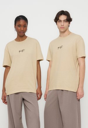 Dvaja mladí dospelí stoja vedľa seba, oblečení v béžových tričkách s malým čiernym nápisovým logom a voľných taupe nohaviciach na jednoliatom pozadí.