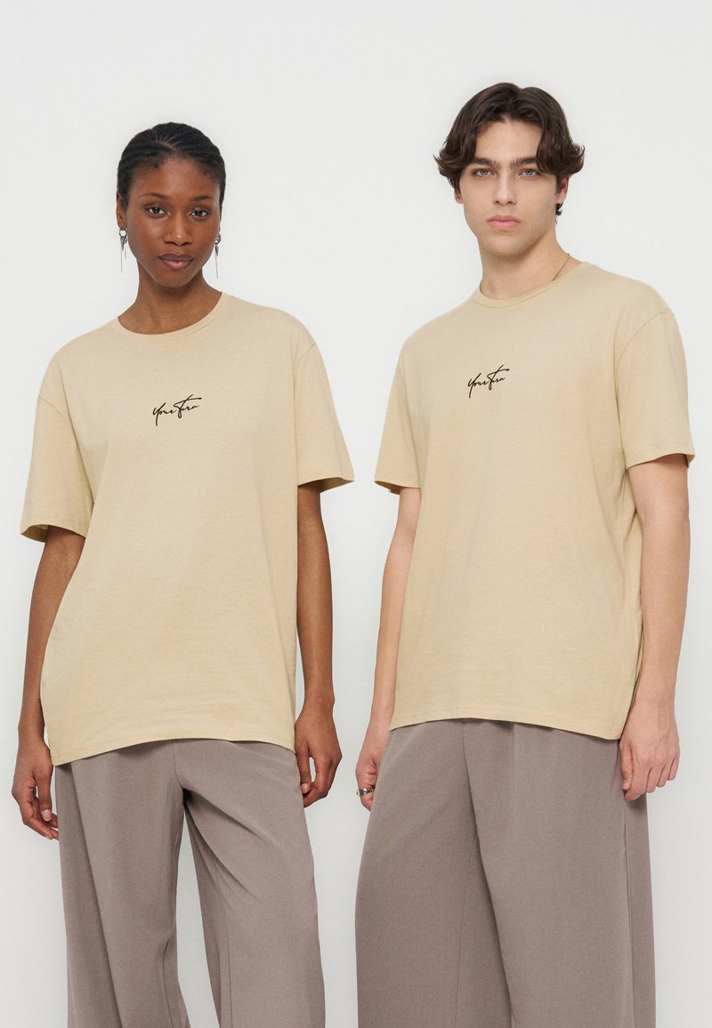 Deux jeunes adultes se tiennent côte à côte, portant des t-shirts beiges avec un petit logo noir en écriture manuscrite et des pantalons taupe amples, sur un fond uni.