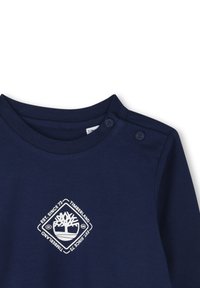 Marineblauw langemouwenshirt met een ronde hals en twee-knopen detail. Heeft een wit logo in een diamantvorm op de borst.