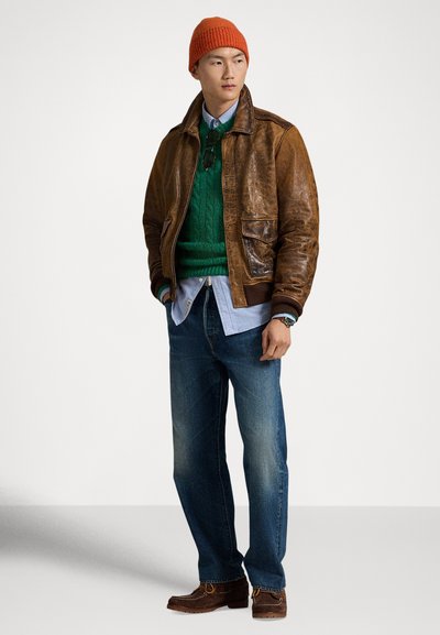 Καφέ δερμάτινο μπουφάν τύπου βομβιστή πάνω από πράσινο πουλόβερ με ανάγλυφο πλέξιμο, ανοιχτό μπλε πουκάμισο, μπλε τζιν και καφέ casual παπούτσια, ολοκληρωμένο με πορτοκαλί πλεκτό σκουφάκι.