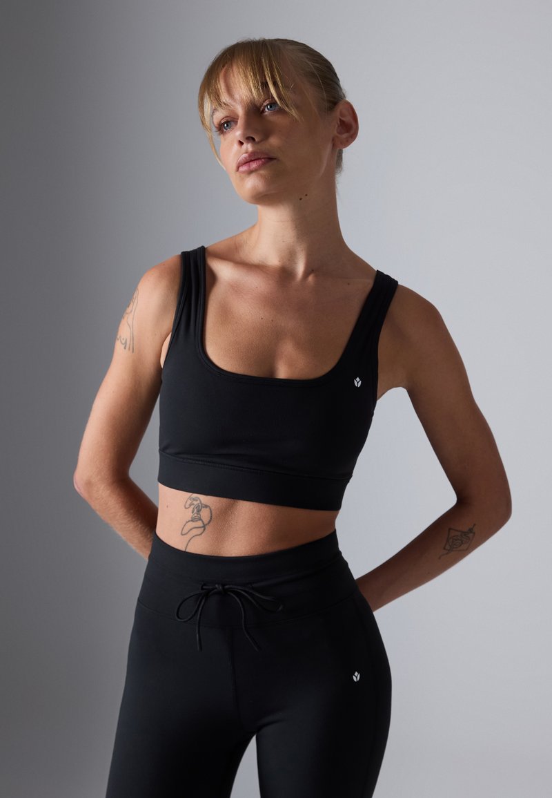 En passerende kvinne med tatoveringer iført svart sports-BH og leggings, står mot en ensfarget grå bakgrunn med hendene bak ryggen.