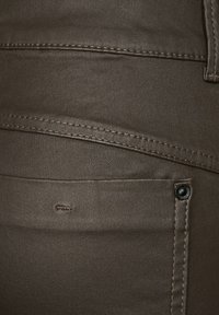 Tmavě hnědé kalhoty s hladkou texturou, viditelným šitím a malým černým tlačítkem na kapse pro detail.